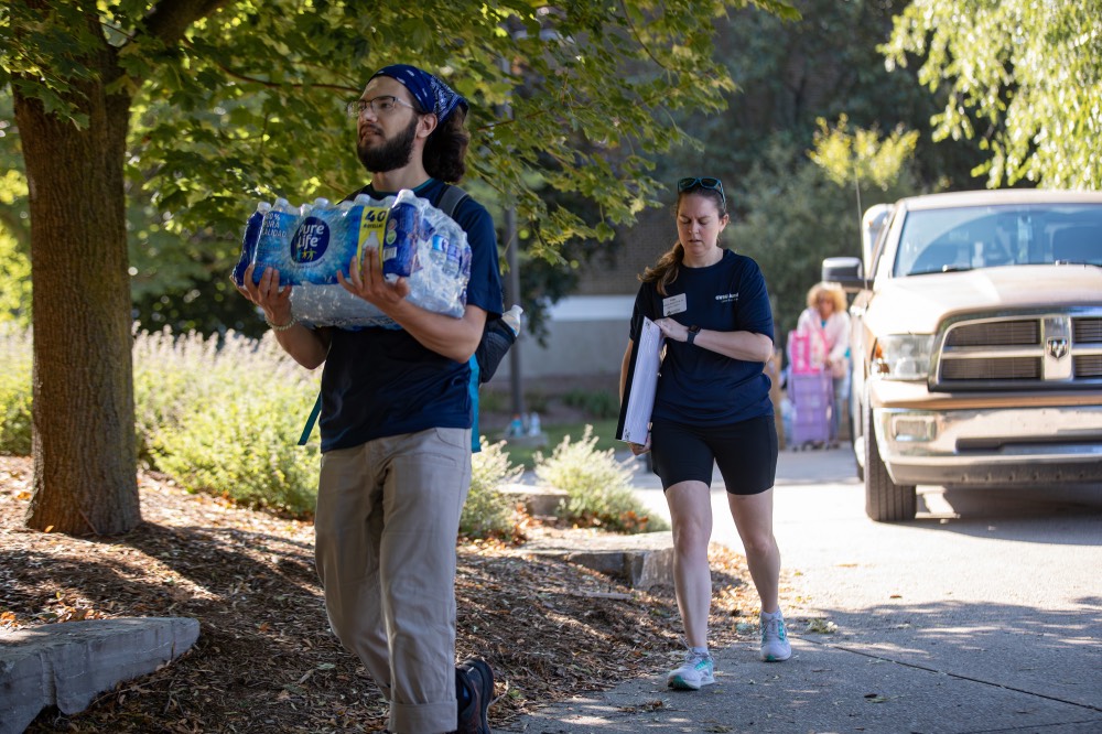 2 GVSU alumni carrying a case of water bottles and a mirror into the dorm building